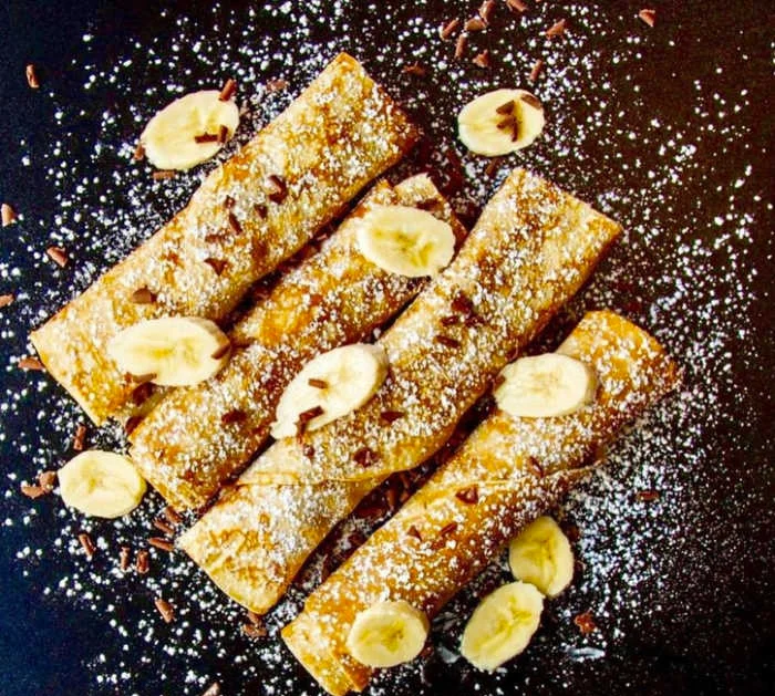 Pancakes on a dark background with bananans sliced on the top and sprinkled with icing sugar.