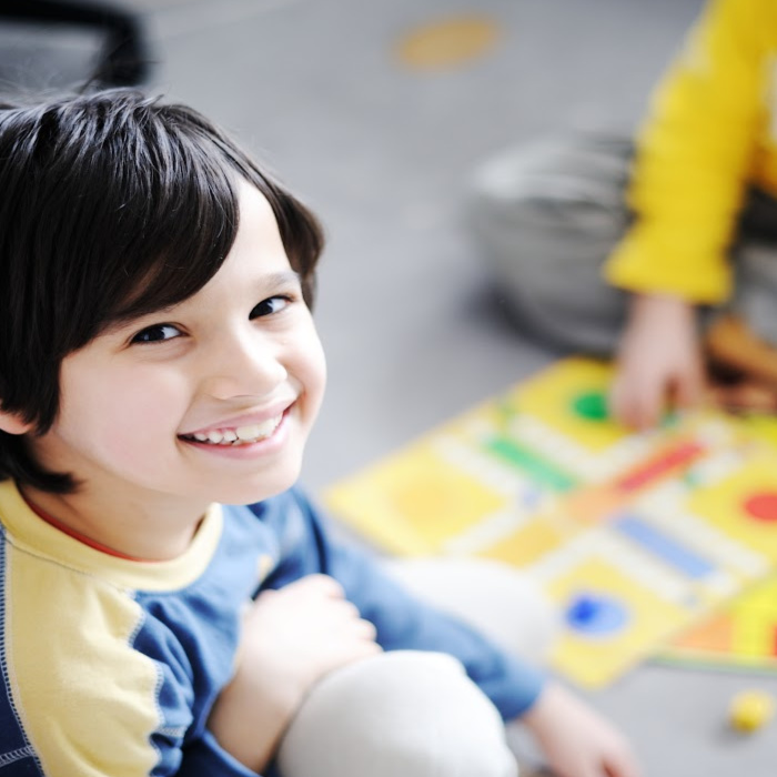 Child smiling at the camera with anoner child playing a board game blurring into the background. 
