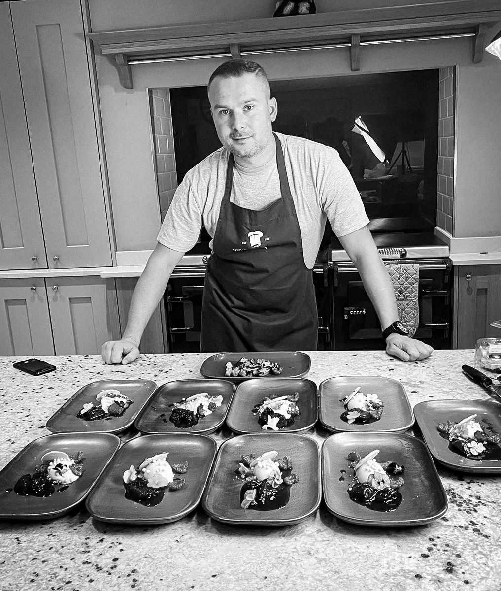 Chef Gourmand wearing an apron stood behind plates with food on, ready to be taken to tables.