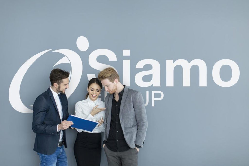3 smartly dressed people, 2 male, 1 female stood infront of light blue/grey wall with a large Siamo logo on it. The people are looking at some paperwork and conversing.