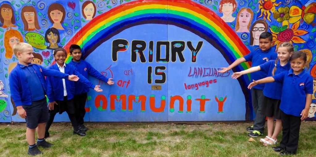Outside wall painted with colourful mural. The painting features a rainbow and the words Priory is Community. In the foreground uniformed students are stood either side of the wall arms gesturing to it.