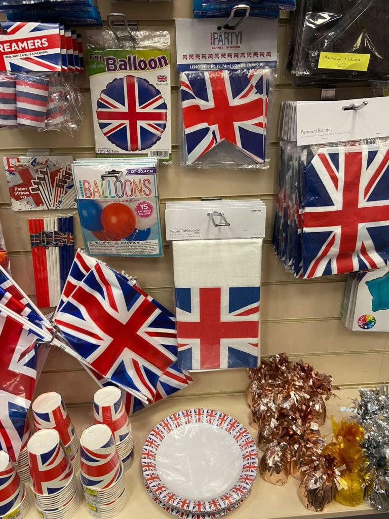 Variety of British themed stock including Union Jack flags, Union Jack plates,napkins,paper cups. some displayed on a lower shelf, some in plastic hang bags above.