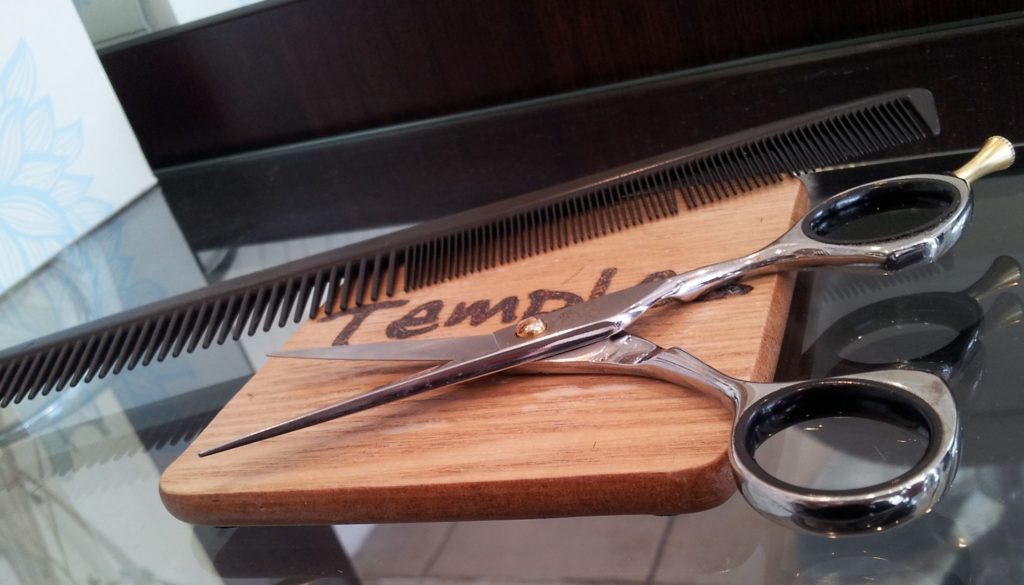 hairdressing scissors and comb resting on a wooden mat with the temples logo on a glass table top.