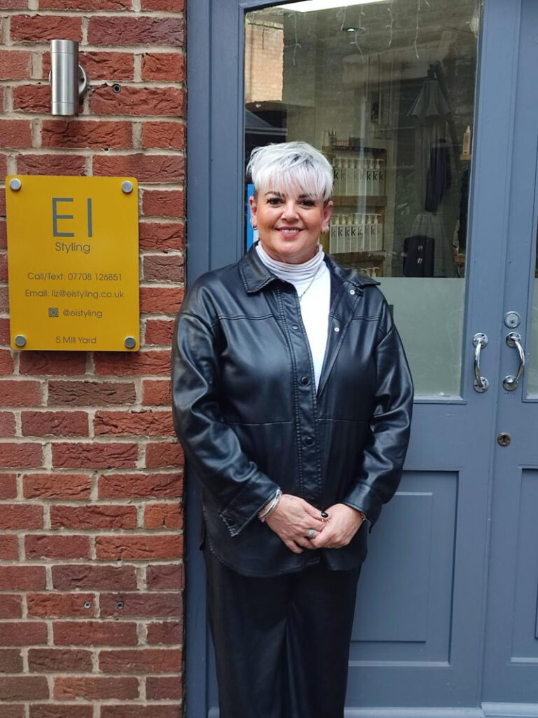 Lady with white hair wearing a black leather jacket in front of a grey door.