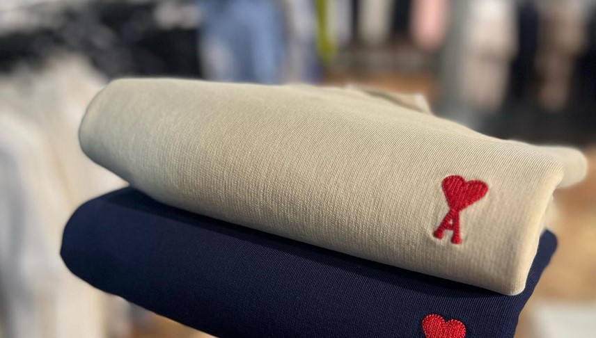 Two sweatshirts on top of each other. One navy blue and the other white. Both have a heart logo in read with the letter A on the breast area.