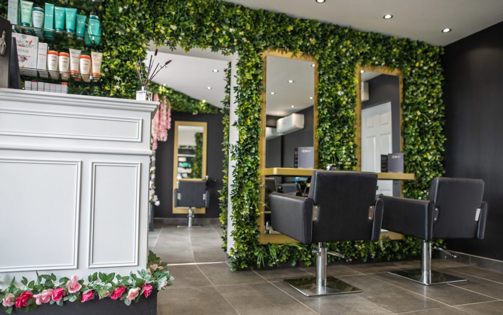 Internal shot of 2 individual leather chairs facing mirrors in front of them hung on a wall covered in green plants.