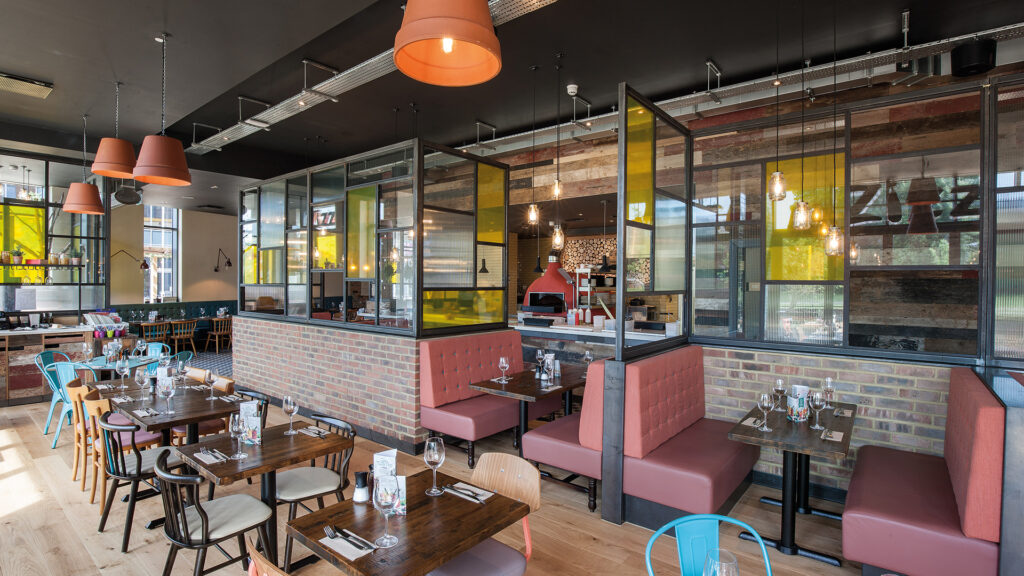 Zizzi interior layout. Multiple wooden tables and chairs in a row. Private leather seated booths with glass wall partitions
