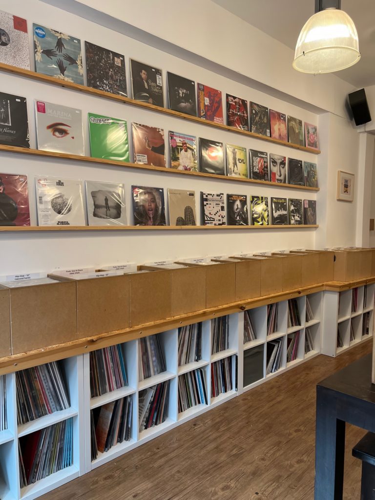 Interior wall with various record covers on 3 shelves running the length of the wall. Below boxed spaces with a number of records in each