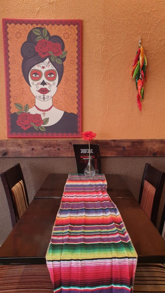 Wooden table and chairs with bright multicolour stripes. Behind is an orange wall with a Dia de los Muertos style portrait of a woman