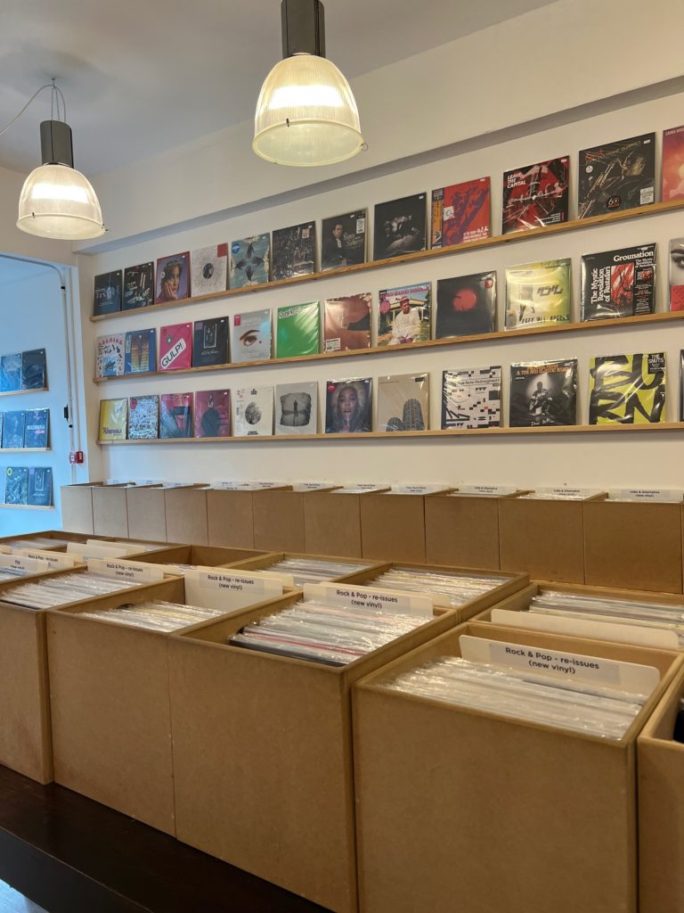 Central table with a number of boxes full of a variety of records. In the background 3 wall shelves displaying records