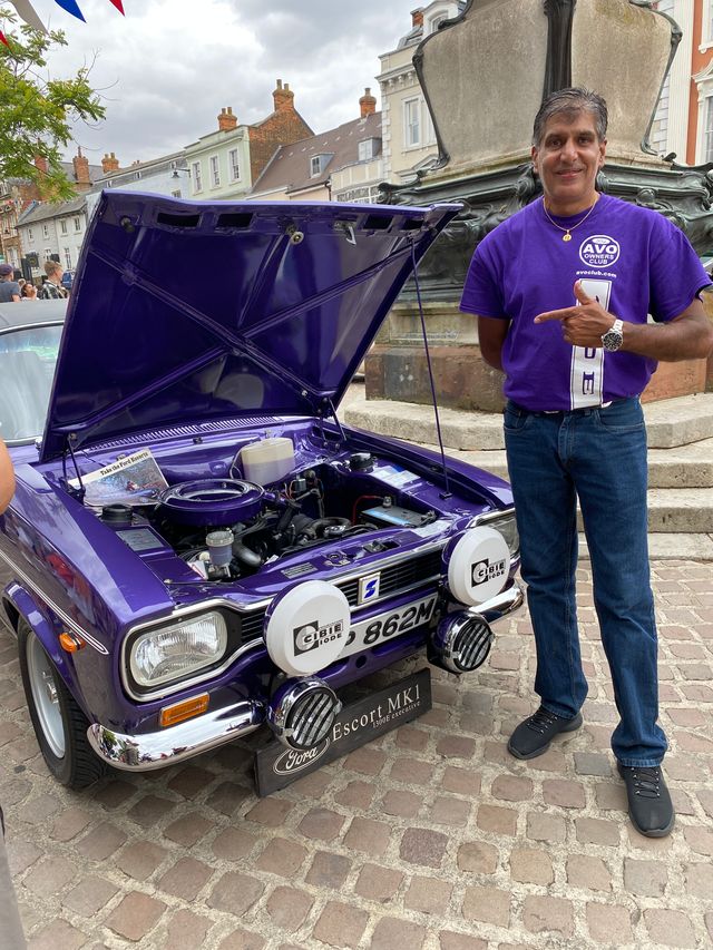 man in a purple t shirt stood next to a purple car with the bonnet opened.