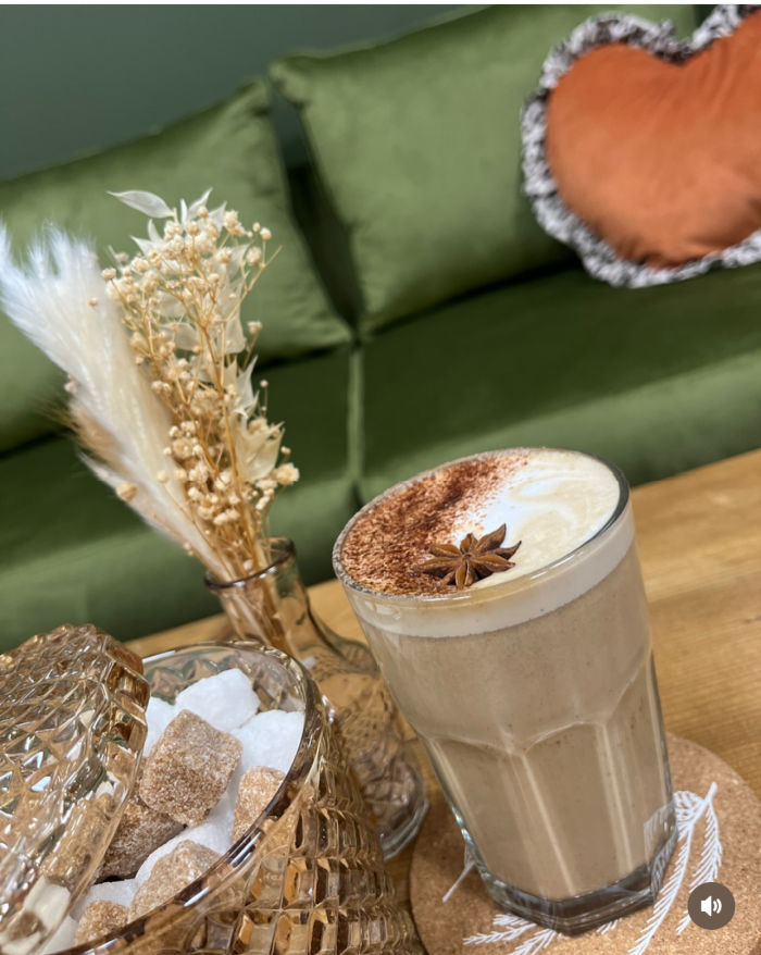 Chai latte in a tall glass next to a bowl of sugar lumps and some dried grasses in a vase.